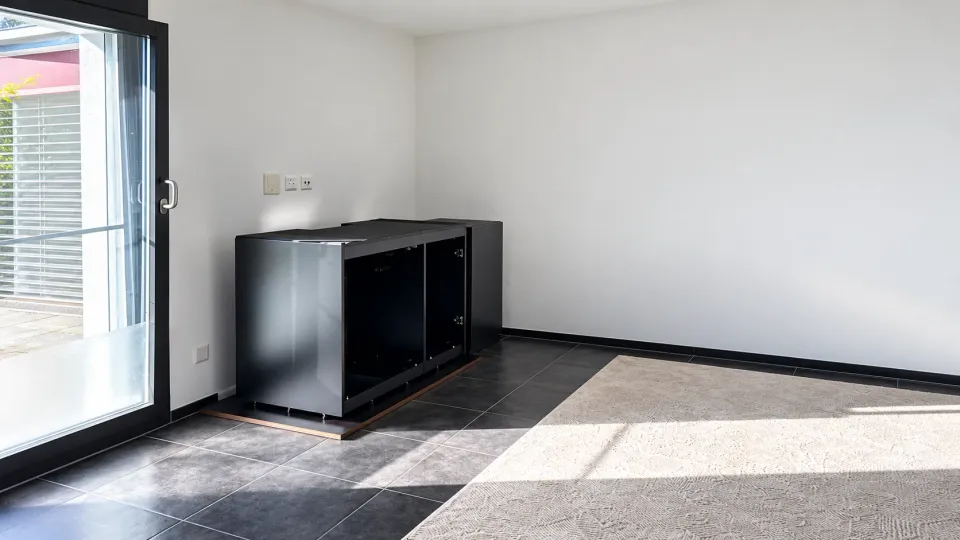 Der schwarze Unterschrank steht noch ohne Becken an der weissen Wand neben der Terrassentür, darunter eine dunkle Platte mit Holzkante. Durch die offene Front sieht man in die leeren Fächer, rechts schliesst der Beistellschrank an. Sonne fällt über den Fliesenboden und den hellen Teppich.