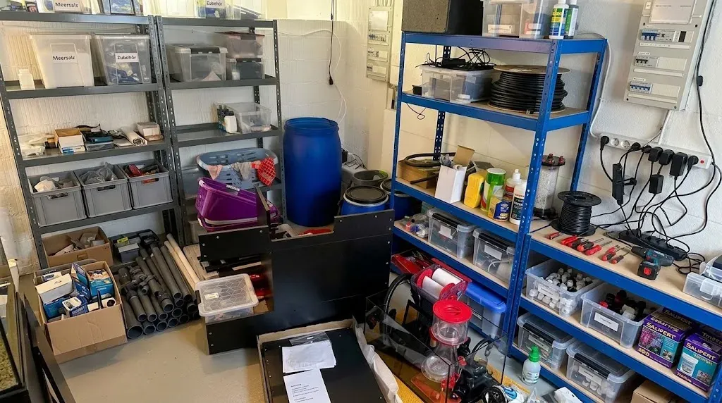 Die Ecke der Waschküche, vollgestellt mit dem, was vorher im Wohnzimmer stand. Links graue Regale mit beschrifteten Boxen für Meersalz und Zubehör, darunter PVC-Rohre und Kartons. In der Mitte das blaue Fass und davor der halb aufgebaute schwarze Unterschrank. Rechts ein blaues Regal mit Schlauchrollen, Werkzeug und Tests, davor das Unterbecken mit dem roten Bügel des ReefMat und dem roten Deckel des alten Abschäumers.