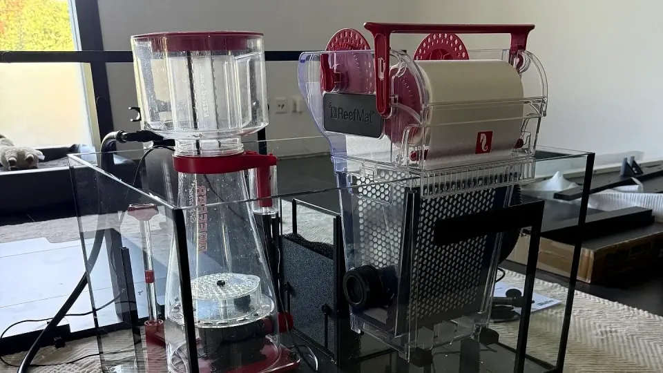Das bestückte Unterbecken steht auf dem Teppich im Wohnzimmer. Links der Reefer Skimmer 600 mit rotem Deckel, rechts der ReefMat 1200 mit der weissen Vliesrolle unter dem roten Bügel. Der ReefMat sitzt quer über der rechten Kammer und belegt gut die Hälfte der Beckenbreite.