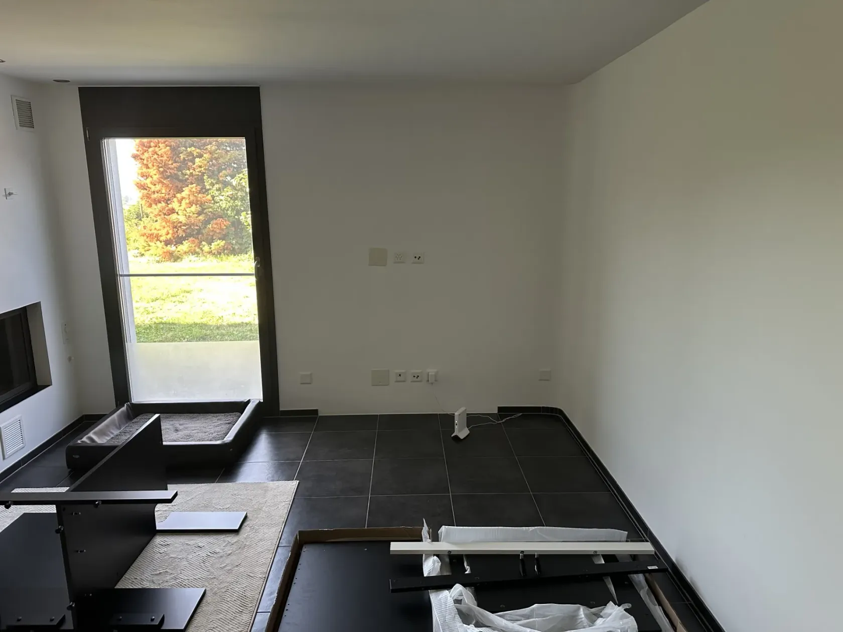 Die Wand ist noch leer, aber davor liegt jetzt der halb aufgebaute Unterschrank — schwarze Seitenteile und Rahmen auf dem Teppich, vorne rechts ein offener Karton mit weiteren Platten, einer Leiste und Folie. Neu steht am Boden vor der Steckdose ein kleines weisses Gerät.